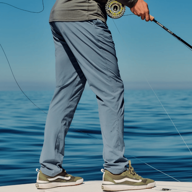 A person is on a boat holding a fishing rod, wearing a long-sleeve shirt, light blue pants, and green-and-white sneakers.