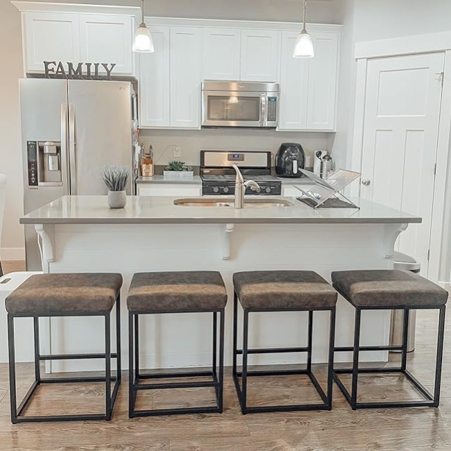 A modern kitchen setup features a sleek white island with a sink, surrounded by four square cushioned bar stools with black metal frames. Stainless steel appliances, including a fridge and oven, complement the minimalist decor.