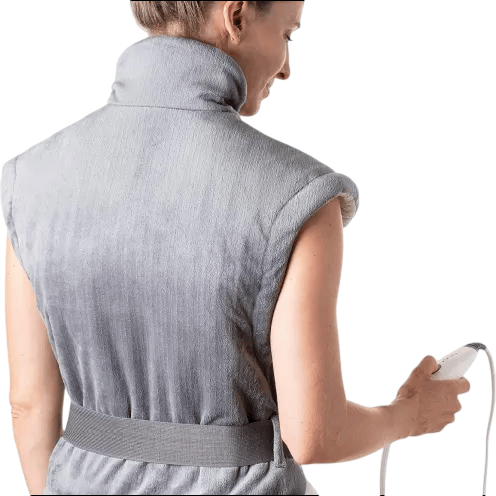 A person wearing a grey electric heating pad with a high collar, designed for neck and shoulder relief, holding a connected control device.