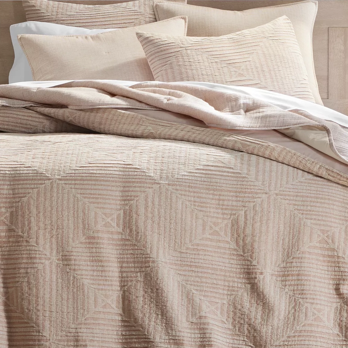 A beige bedding set with textured geometric patterns, including a quilt or comforter and matching pillow shams, is displayed neatly arranged on a bed.