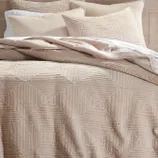A beige bedding set with textured geometric patterns, including a quilt or comforter and matching pillow shams, is displayed neatly arranged on a bed.