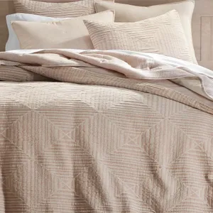 A beige bedding set with textured geometric patterns, including a quilt or comforter and matching pillow shams, is displayed neatly arranged on a bed.