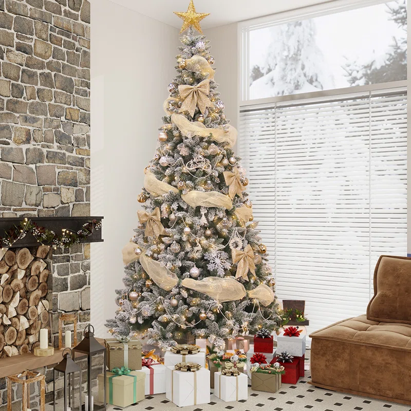 A snow-dusted Christmas tree decorated with gold ribbons, bows, and ornaments, topped with a star. Surrounded by wrapped presents, it stands next to a fireplace with visible logs and lanterns. Large windows reveal a snowy outdoor scene.