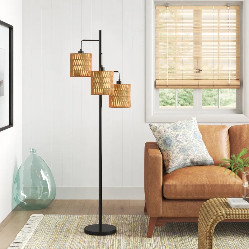 A modern floor lamp with three woven rattan shades stands next to a brown leather sofa with a floral pillow, set in a cozy living room with a light-colored rug and bamboo blinds.