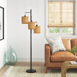 A modern floor lamp with three woven rattan shades stands next to a brown leather sofa with a floral pillow, set in a cozy living room with a light-colored rug and bamboo blinds.