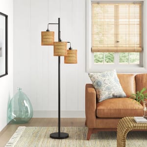 A modern floor lamp with three woven rattan shades stands next to a brown leather sofa with a floral pillow, set in a cozy living room with a light-colored rug and bamboo blinds.
