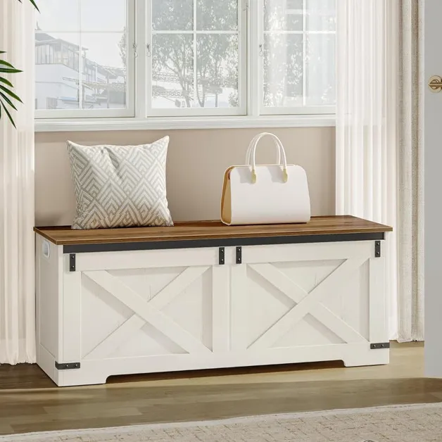 A wooden storage bench with a white base and dark wood top is placed under a window, featuring a geometric-patterned cushion and a white handbag on top. Slippers rest on the floor nearby.