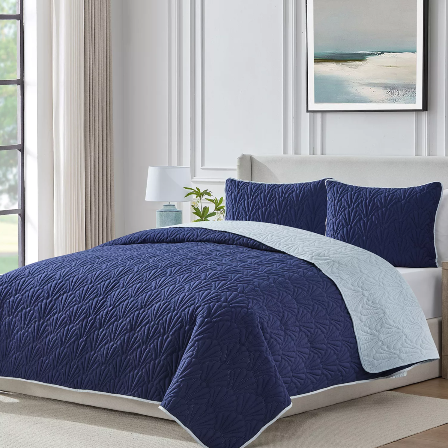 A blue quilted bedding set with a leaf pattern, including a comforter and matching pillow shams, is displayed in a modern bedroom with an abstract painting on the wall and a bedside lamp.