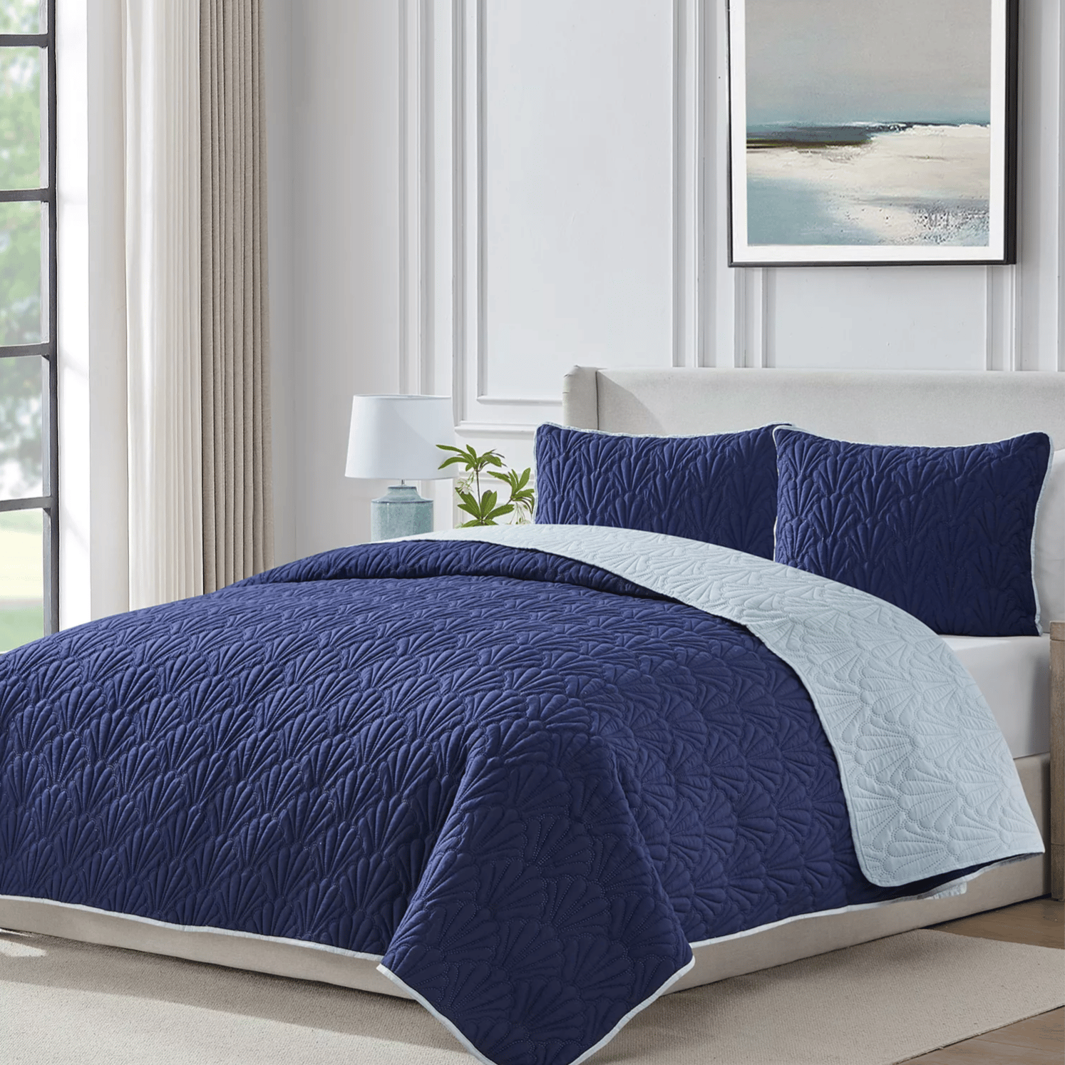 A blue quilted bedding set with a leaf pattern, including a comforter and matching pillow shams, is displayed in a modern bedroom with an abstract painting on the wall and a bedside lamp.