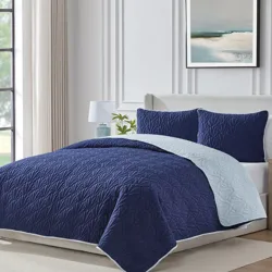A blue quilted bedding set with a leaf pattern, including a comforter and matching pillow shams, is displayed in a modern bedroom with an abstract painting on the wall and a bedside lamp.
