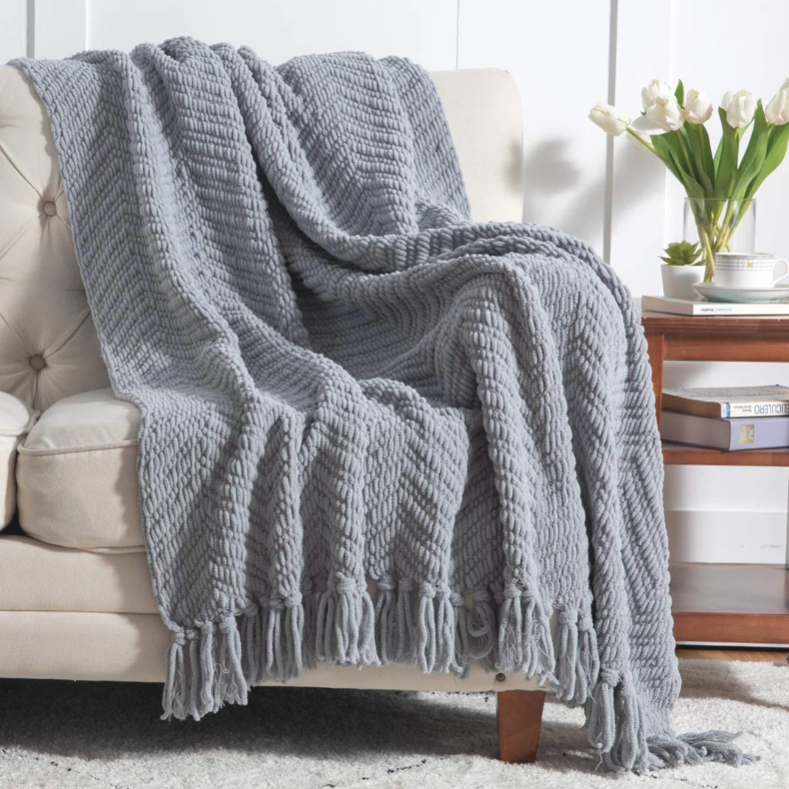 A gray knitted throw blanket with fringe is draped over a beige couch. Nearby, a wooden side table holds a vase of white tulips, a cup, and stacked books.