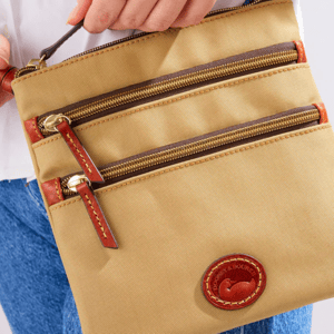 A tan Dooney & Bourke crossbody bag with two front zippered pockets, leather trim, and gold-tone hardware, featuring the brand's embossed logo on a circular leather patch.