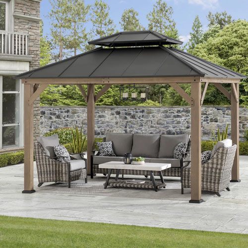 A gazebo with a black metal roof and wooden frame, featuring outdoor wicker furniture and a central coffee table, situated on a patio with a stone wall and garden backdrop.