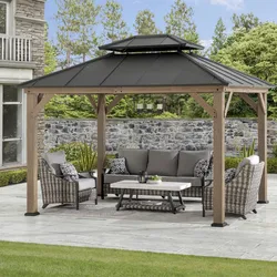 A gazebo with a black metal roof and wooden frame, featuring outdoor wicker furniture and a central coffee table, situated on a patio with a stone wall and garden backdrop.