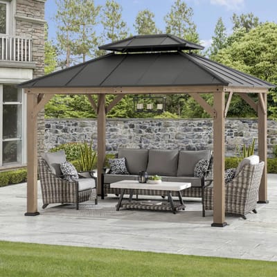 A gazebo with a black metal roof and wooden frame, featuring outdoor wicker furniture and a central coffee table, situated on a patio with a stone wall and garden backdrop.