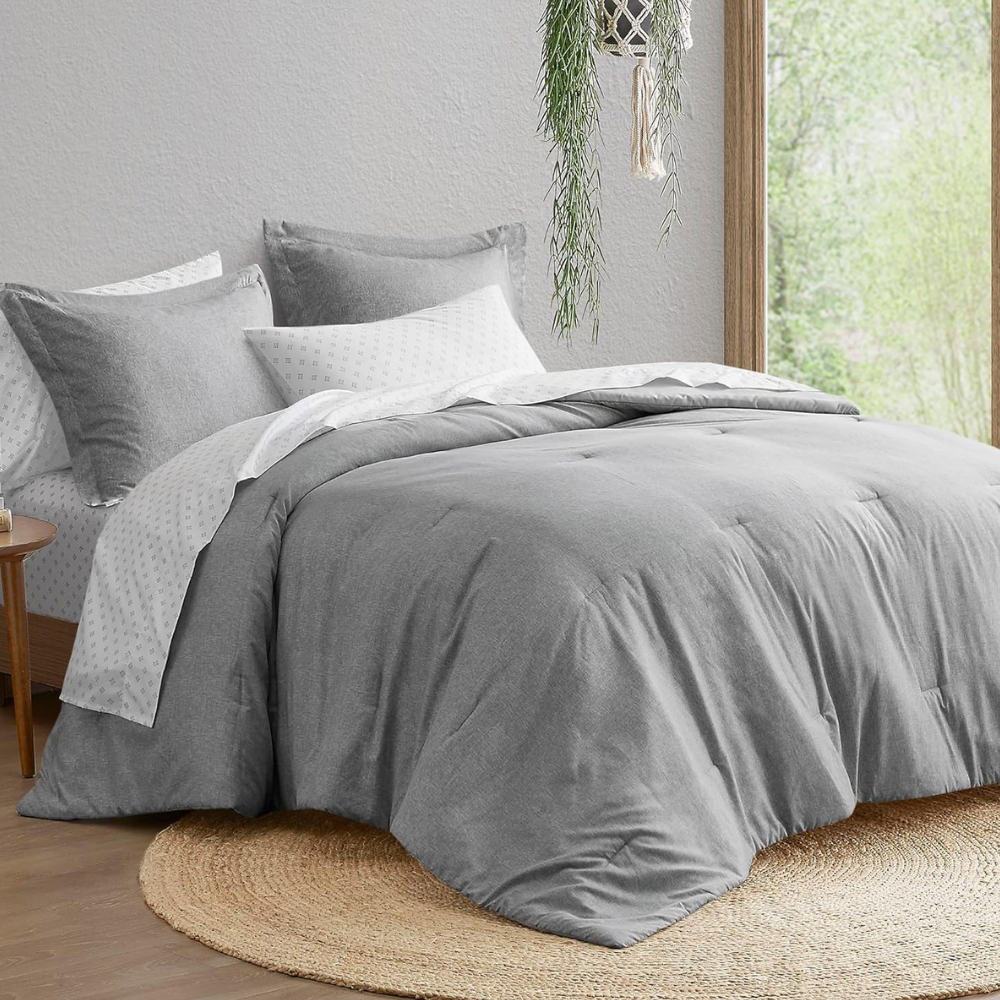 A neatly arranged bed with a gray comforter and pillows, paired with a white sheet featuring a subtle pattern. The setting includes a round jute rug and a hanging plant near a window.