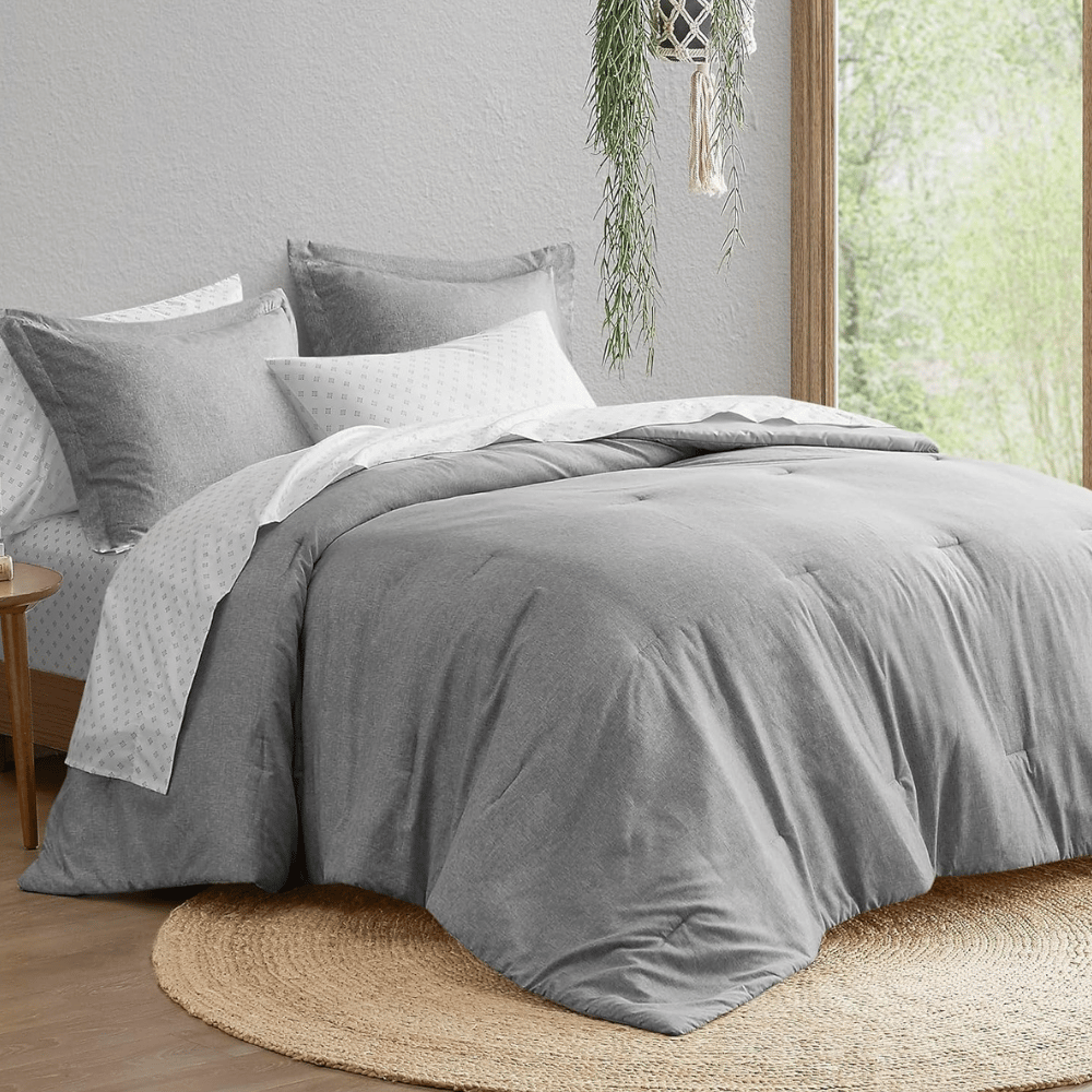 A neatly arranged bed with a gray comforter and pillows, paired with a white sheet featuring a subtle pattern. The setting includes a round jute rug and a hanging plant near a window.