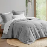 A neatly arranged bed with a gray comforter and pillows, paired with a white sheet featuring a subtle pattern. The setting includes a round jute rug and a hanging plant near a window.