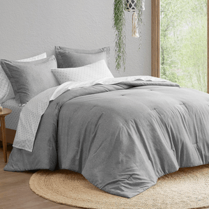 A neatly arranged bed with a gray comforter and pillows, paired with a white sheet featuring a subtle pattern. The setting includes a round jute rug and a hanging plant near a window.