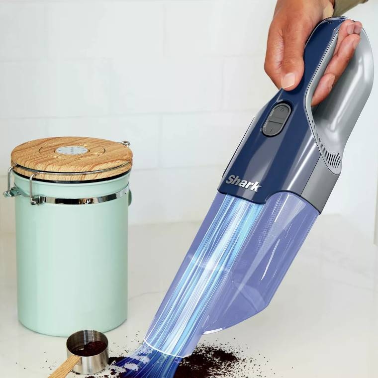 A Shark handheld vacuum cleaner is being used to pick up a spill, showing its suction power and compact size.