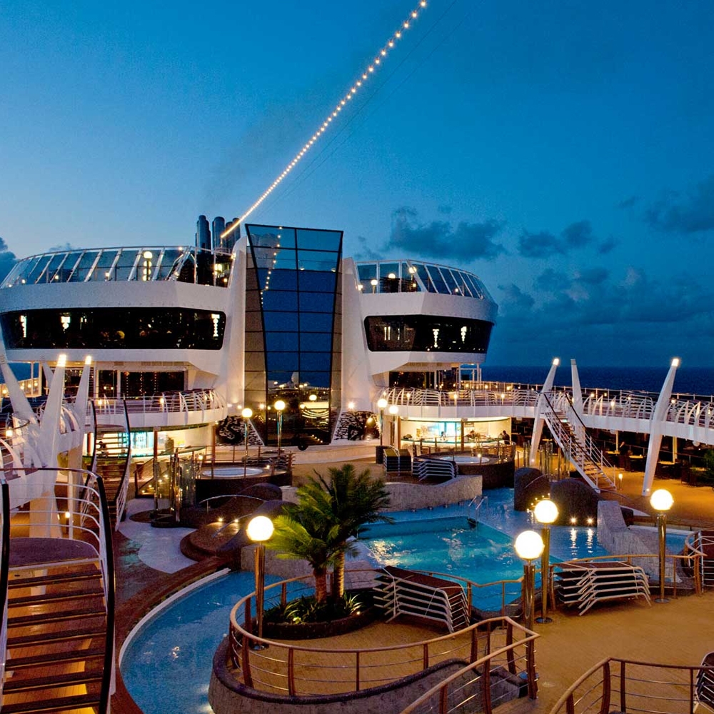 A luxury cruise ship deck features a swimming pool, modern architecture, and ambient lighting under a twilight sky, creating a relaxing atmosphere for passengers.