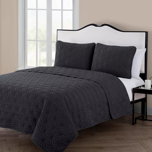 A charcoal gray quilt and pillow shams set with a geometric pattern, displayed on a bed with a contrasting white and black headboard, accompanied by a small side table.