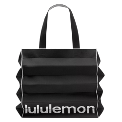 A black pleated tote bag with white piping and the \“lululemon\“ logo in white at the bottom.
