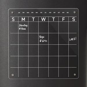 Magnetic acrylic calendar with a weekly grid layout is shown on a stainless steel surface. It displays notes such as \“Meeting @ Noon,\“ \“Yoga @ 6PM,\“ and \“Lake!\“ in white marker.