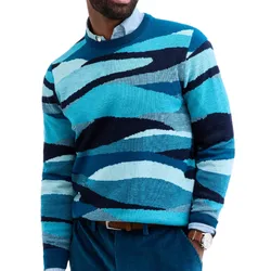 A man wears a multicolored, abstract-patterned sweater in shades of blue, paired with a light blue shirt, teal pants, and a brown belt.
