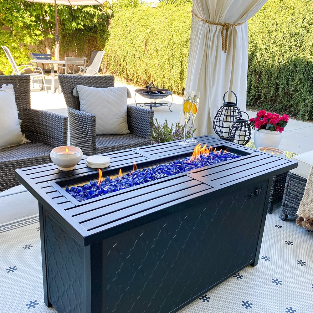 A rectangular outdoor gas fire pit with blue glass rocks on a patio, surrounded by wicker furniture and decorative items.