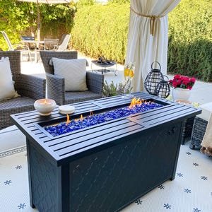 A rectangular outdoor gas fire pit with blue glass rocks on a patio, surrounded by wicker furniture and decorative items.