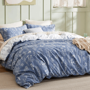 A bed is made with blue and white floral patterned bedding, including a comforter, pillowcases, and a throw blanket, set in a room with light green walls and natural lighting.