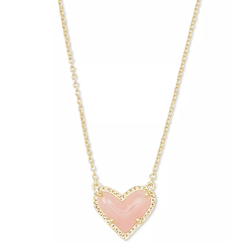 A Kendra Scott necklace featuring a heart-shaped pendant with a soft pink stone set in a gold frame. The chain is delicate and gold-toned, complementing the elegant design.