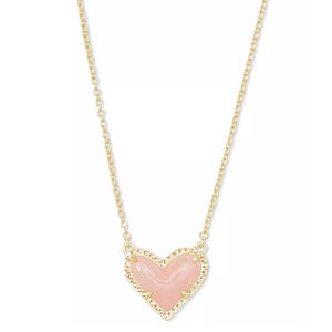 A Kendra Scott necklace featuring a heart-shaped pendant with a soft pink stone set in a gold frame. The chain is delicate and gold-toned, complementing the elegant design.