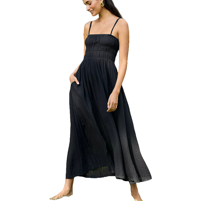 A woman is wearing a black maxi dress featuring thin straps, a smocked bodice, and a flowy, ankle-length skirt. She accessorizes with large hoop earrings and is barefoot.