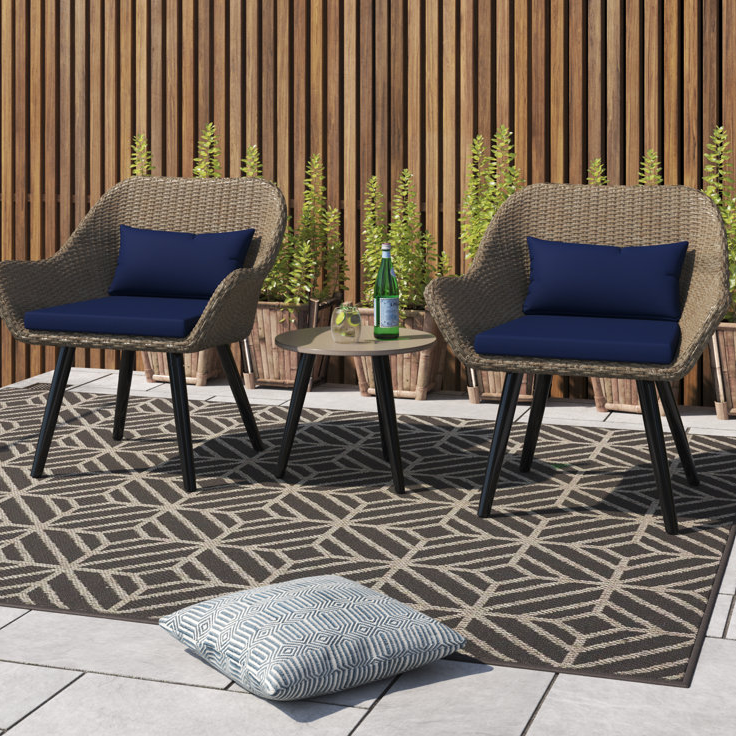 A 3-piece patio set featuring two wicker chairs with dark blue cushions and a small round table sits on a patterned area rug. It is complemented by a wooden slat fence and potted plants in the background.