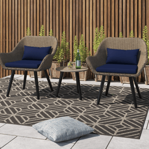 A 3-piece patio set featuring two wicker chairs with dark blue cushions and a small round table sits on a patterned area rug. It is complemented by a wooden slat fence and potted plants in the background.