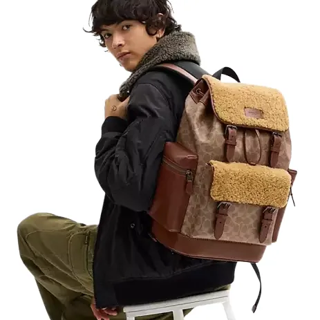 A stylish brown backpack with a fleece flap and leather accents is worn by a person in a black jacket with a shearling collar and green pants.