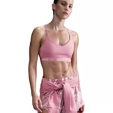 Nike Women's Indy Sports Bra in pink, featuring thin straps and a supportive elastic band with the Nike logo. Paired with matching patterned shorts and an additional shirt tied around the waist.