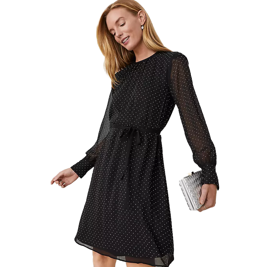 A woman is wearing a black dress with small white polka dots, featuring long sheer sleeves and a tie at the waist. She holds a small metallic clutch.