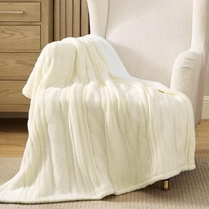 A soft, cream-colored throw blanket with a dual-texture design featuring a cable-knit pattern on one side and plush sherpa on the other, draped over a cozy, light-colored armchair with gold-tipped feet.