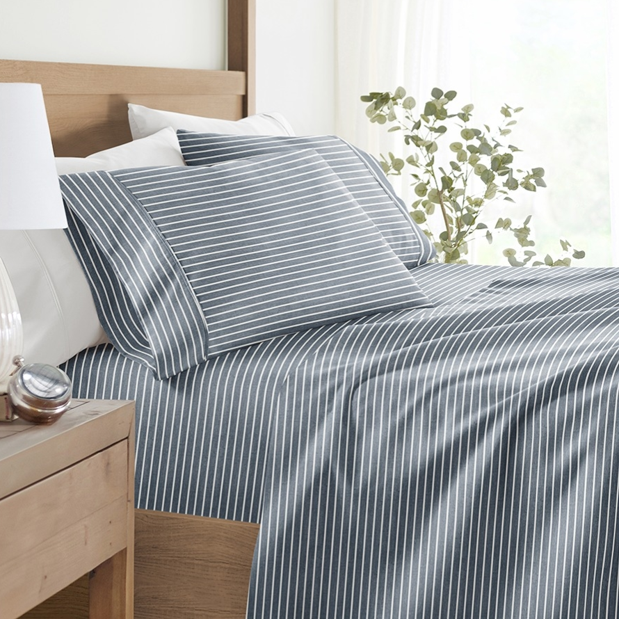 A neatly made bed features blue and white striped bedding, accompanied by a bedside table with a white lamp, a small succulent plant, and a clock.