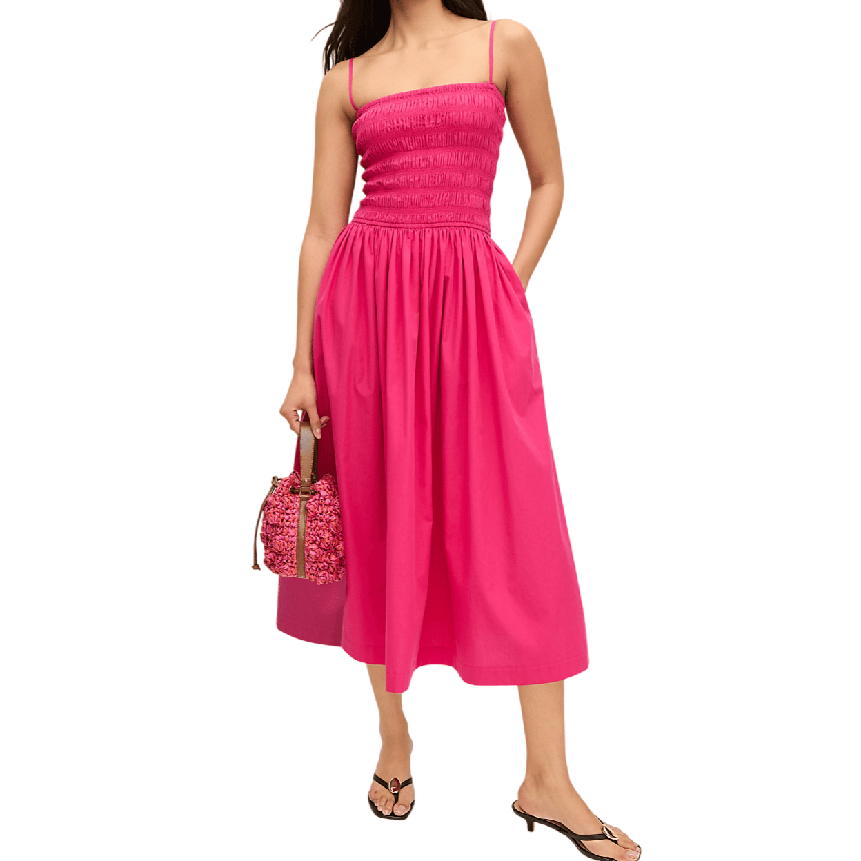 A woman wears a bright pink midi sundress with a smocked bodice and thin straps, paired with black sandals. She carries a pink textured handbag.