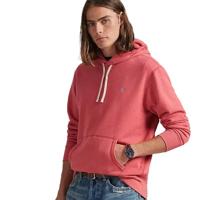 A person is wearing a pink hoodie with a small embroidered logo on the chest, paired with blue jeans and a watch.