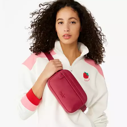 A woman is wearing a white quarter-zip pullover with pink accents and a small embroidered fruit design, paired with a red crossbody bag.