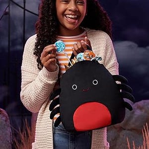 A smiling person is holding a black plush Halloween pail with an orange interior and a simple face design, resembling a spider with six soft, striped legs.