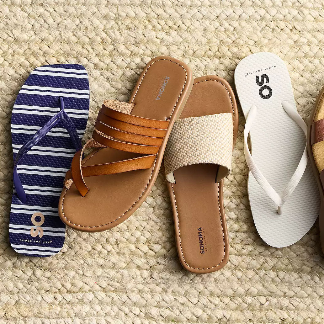 Four styles of sandals on woven fabric: a blue and white striped flip-flop, a brown multi-strap leather slide, a beige woven slide, and a white flip-flop.