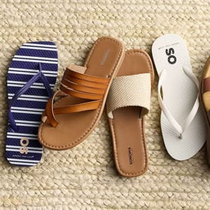 Four styles of sandals on woven fabric: a blue and white striped flip-flop, a brown multi-strap leather slide, a beige woven slide, and a white flip-flop.