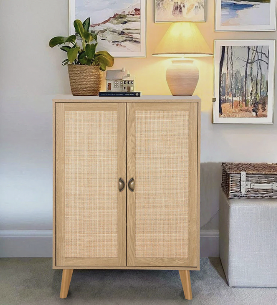 A 43\“ rattan cabinet with a natural wooden frame and two cane-paneled doors, featuring dual metal handles. Above it sit a lamp, a small plant in a woven basket, and decorative items. The setting includes framed artwork on the wall.