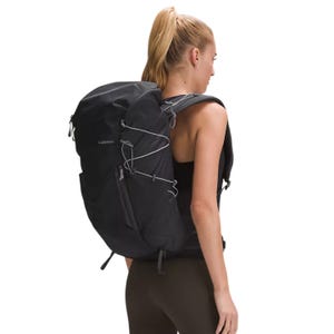 A person wearing a black lululemon 28L Hiking Backpack with a sleek design, featuring adjustable straps and external bungee cords for extra storage. The backpack is worn by a female model in athletic attire.